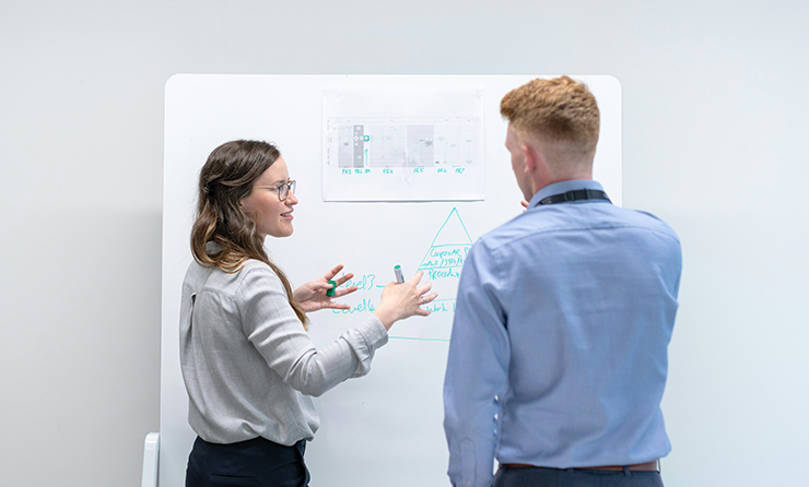 En man och en kvinna står med ryggen mot kameran och diskuterar något vid en whiteboard-tavla.
