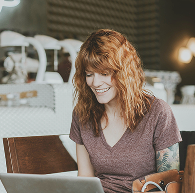 En rödhårig kvinna i t-shirt med synlig tatuerad arm sitter på ett café eller ett kontor och skriver på sin laptop.