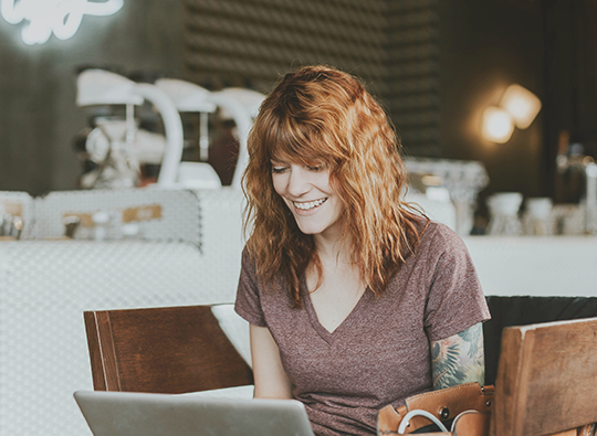 En rödhårig kvinna i t-shirt med synlig tatuerad arm sitter på ett café eller ett kontor och skriver på sin laptop.
