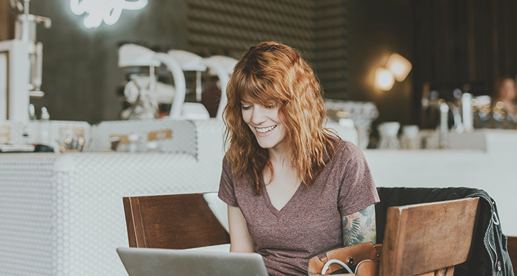 En rödhårig kvinna i t-shirt med synlig tatuerad arm sitter på ett café eller ett kontor och skriver på sin laptop.