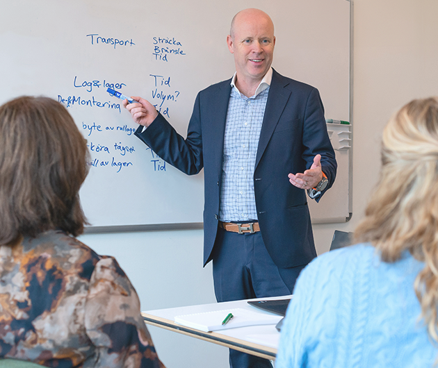 En man i skjorta och kostym står vid en whiteboard-tavla och pekar/pratar för ett gäng personer i ett konferensrum.