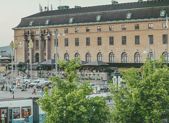 Vy över Göteborgs Centralstation med Clarion Post hotel i bakgrunden. Spårvagnar och gröna lummiga träd syns.