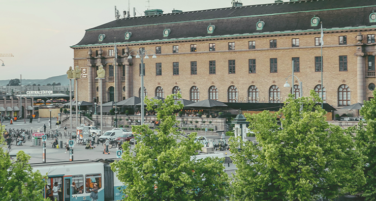 Vy över Göteborgs Centralstation med Clarion Post hotel i bakgrunden. Spårvagnar och gröna lummiga träd syns.